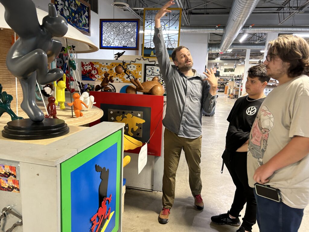 A man is gesticulating with his arms, as he faces two teenagers who are looking at some sculptures. The man holds one arm high and curving over his head and the other is folded with fingers clutched. The is speaking to the students, probably explaining the significance of the sculpture they are viewing, which is a group of small abstract human figures.