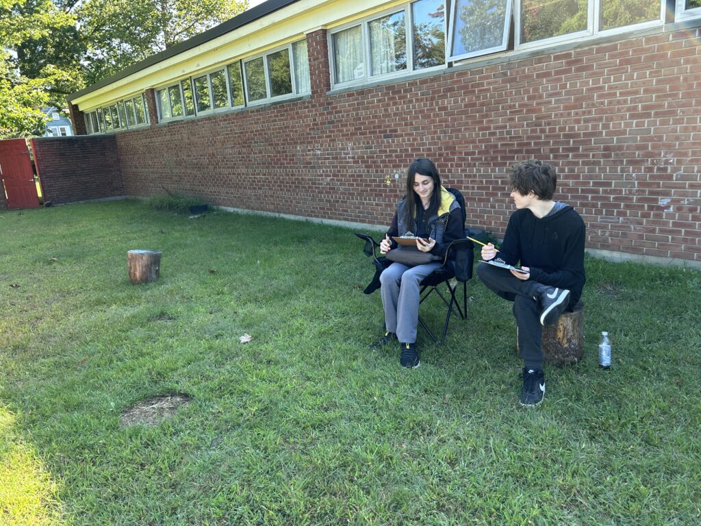 Two teenaged boys sitting in camp chairs on a grassy lawn with a brick wall behind them. They are holding clipboards and chatting in a friendly way.