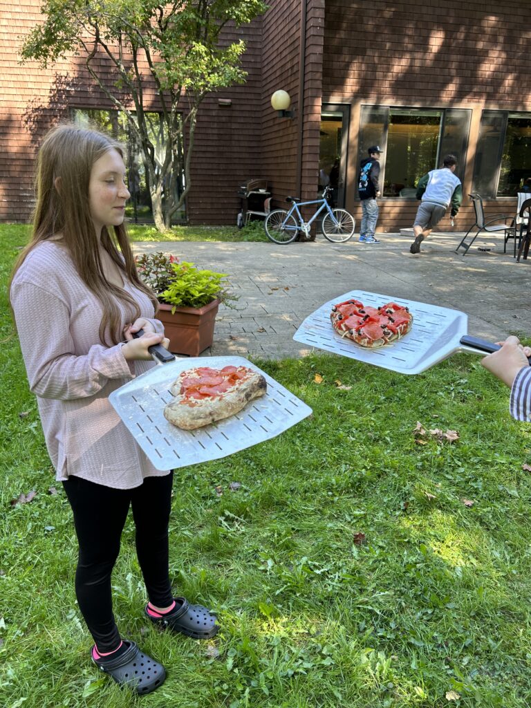 A smiling teenager holds a pizza on a long-handled pizza peel. Another peel with a pizza is at the right of the photo, but the person holding it is not in the frame.