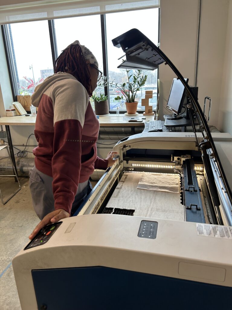 A student is leaning on and looking into a large boxy machine with the lid raised. The machine contains a flat piece of wood and the student is examining the wood. This is a laser cutting tool and she is determining whether the wood is aligned correctly.