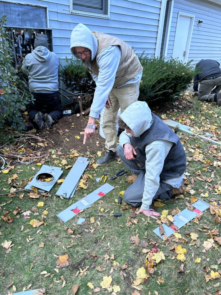 A man and a younger man, both wearing hooded sweatshirts with the hoods up, examine some metal parts on a leaf-covered lawn as a third man behind them works on an electrical panel attached to the side of a house.