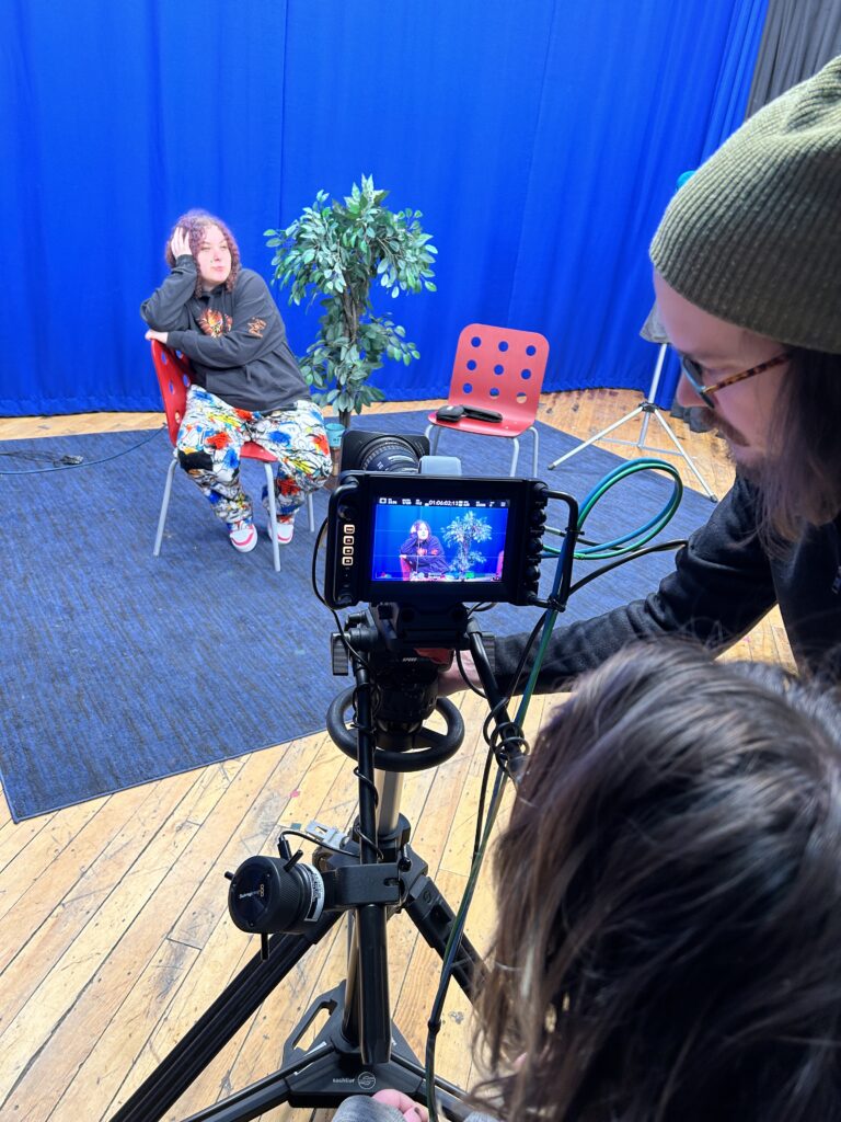 A man shows someone how to use a video camera in a TV studio whlle a person sitting in a chair in the background seems to be bored, waiting for the show to begin.