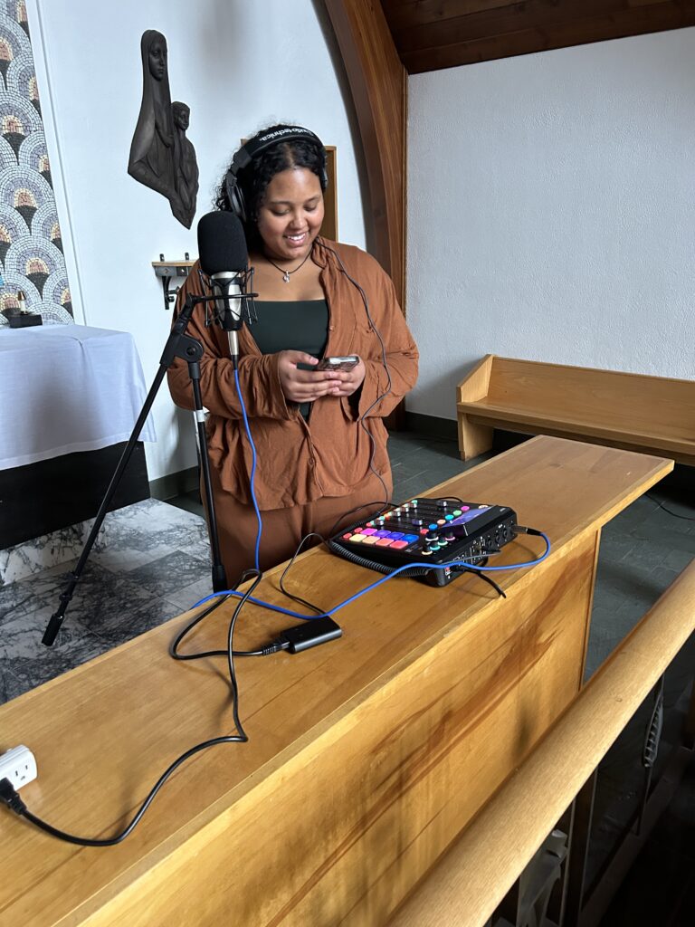 Teenager in a chapel with recording equipment in the foreground. She is working with the equipment as if getting ready to record, and she has a smile on her face and looks happy.