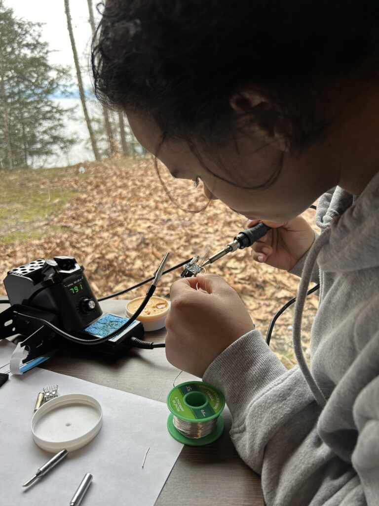 Teenage boy soldering next to a window. Fall leaves and a lake are in the background.