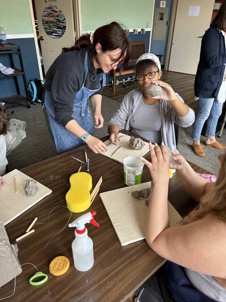 Standing woman in apron helps a student sitting at a table with a pottery project. The student is holding up a spherical piece of pottery to the camera to show the viewer.