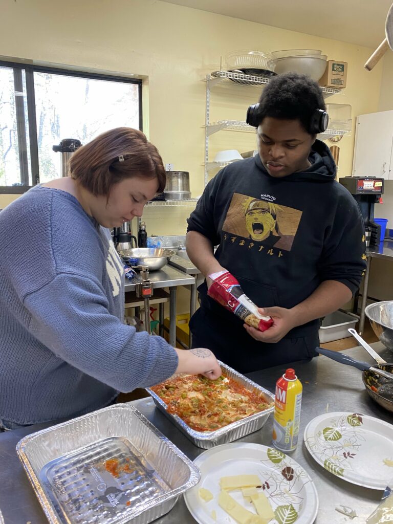 Two teenagers are sprinkling shredded cheese on top of a casserole in a large aluminum tray.