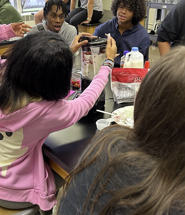 Students watching expectantly while a girl puts the lid on a blender.