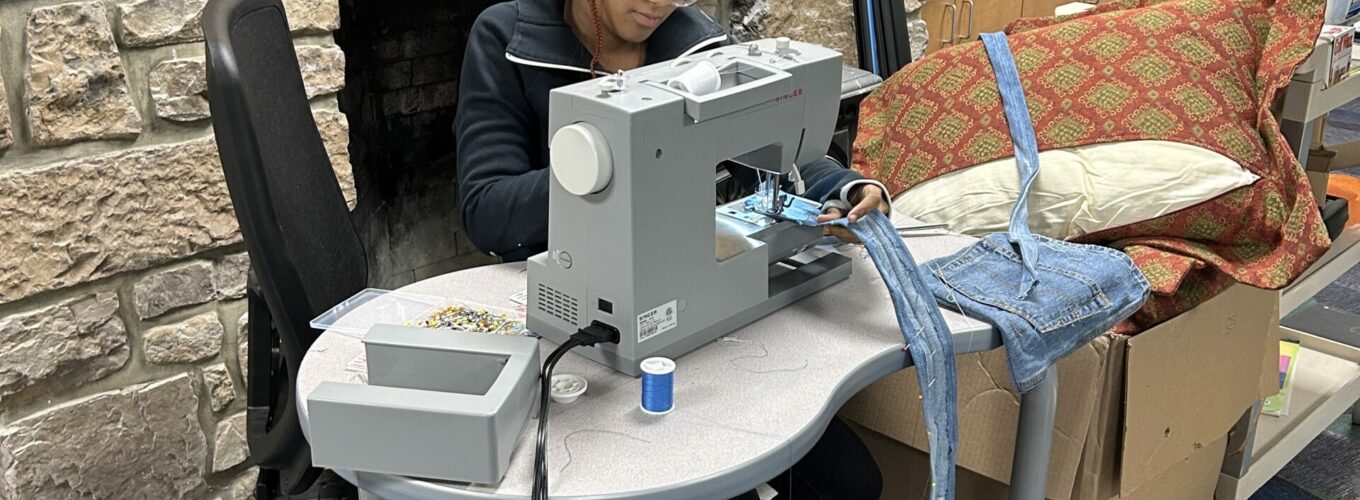 Student working at a sewing machine with stone fireplace hearth in background.