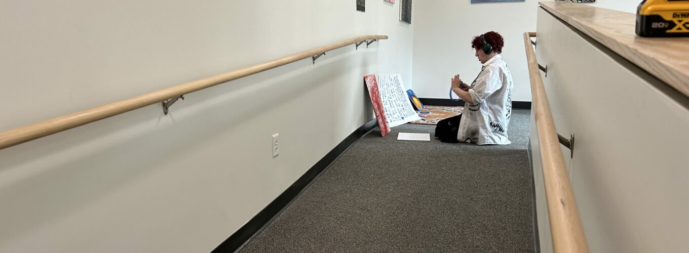 Student at the end of a ramp prepares artwork for hanging in galleriy. Artwork hangs on the wall behind them.